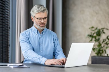 Modern ofis ortamında dizüstü bilgisayarıyla çalışan olgun bir iş adamı. Gri saçlı ve gözlüklü adam açık mavi bir gömlek giyiyor. Konsantrasyon ve verimlilik taşıyor..