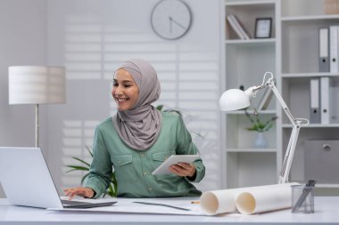 Modern bir ofiste çalışan tesettürlü bir iş kadını. Dizüstü bilgisayar kullanıyor ve elinde bir tablet var. Kendinden emin ve profesyonel görünüyor. Çevre düzenli ve elverişli.