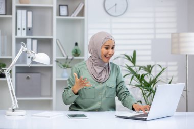 Modern bir ofis ortamında online video görüşmesi yapan tesettürlü mutlu bir kadın. Tablet ve lambayla beyaz bir masada dizüstü bilgisayar kullanırken gülümsüyor ve el kol hareketi yapıyor..