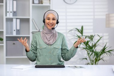Dost canlısı bayan müşteri hizmetleri temsilcisi kulaklık takıyor ve modern ofis ortamında el kol hareketi yaparken gülümsüyor. Profesyonel ve cana yakın, müşterilere yardım etmeye hazır.