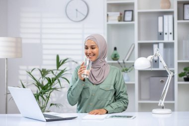 Modern ofis masasında çalışırken tesettürlü gülümseyen kadın biraz su içiyor. Düzenli çalışma alanı dizüstü bilgisayar, lamba ve minimalist dekorla donatılmıştır..