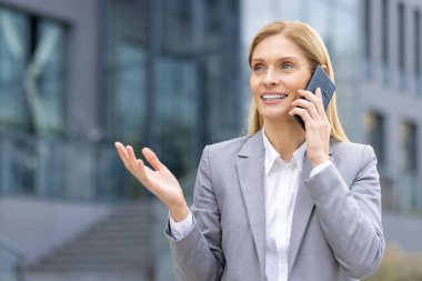 Gri takım elbiseli, kendine güvenen bir iş kadını, modern bir ofis binasının önünde duruyor, bir telefon görüşmesi yapıyor. İşle ilgili konuları konuşurken gülümsüyor ve jestler yapıyor..