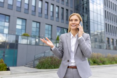 Gri takım elbiseli kendine güvenen bir iş kadını modern ofis binalarının önünde olumlu bir telefon görüşmesi yapıyor. Nişanlı ve mutlu görünüyor.
