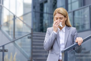 İş elbisesi giymiş profesyonel bir kadın, çağdaş bir ofis binasının önünde mendil tutarken hapşırıyor. Alerji belirtileri gösteriyor. Ya da soğuk algınlığı..