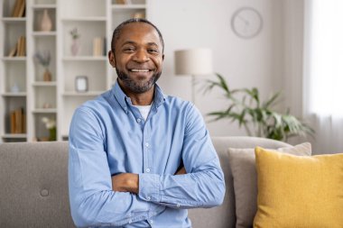 Parlak bir oturma odasında rahat bir kanepede oturan gülümseyen bir adamın portresi. Kollarını kavuşturmuş mavi bir gömlek giyiyor. Arkaplanda bir kitaplık ve rahat bir ev dekorasyonu var..