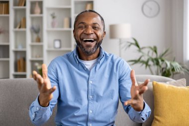 Mavi tişörtlü neşeli bir adam evden keyifli bir video görüşmesi yapıyor. Modern, rahat bir oturma odasında rahat bir koltukta oturuyor, davetkar bir atmosferi var, gülümsüyor ve enerjik bir şekilde el kol hareketi yapıyor..