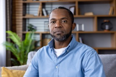 Mavi gömlekli ciddi bir adam modern bir oturma odasında bir kanepede oturuyor. Arkaplanda, rahat ve profesyonel bir atmosfer yaratan ahşap bir kitaplık ve bitki bulunuyor..