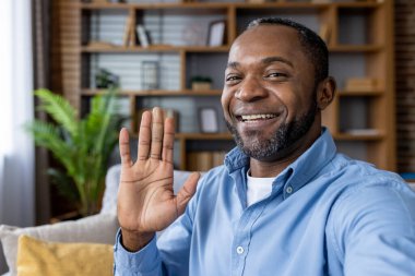 Dostça gülümseyen ve el sallayan bir adam samimi ev ortamından gelen bir video görüşmesi sırasında. Sanal iletişim, dostluk ve uzaktan çalışma kavramları için ideal..