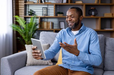 Rahat giysiler içinde gülümseyen adam rahat oturma odasında kanepede otururken tablette arkadaşça görüntülü bir sohbet yapıyor. Görüntü, iletişim ve teknoloji kavramlarını iletir..