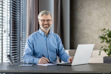 Gözlüklü ve mavi gömlekli olgun bir işadamı gülümsüyor ve modern ofis ortamında dizüstü bilgisayarla çalışıyor. Ofiste panjurlu büyük pencereler ve kırtasiye tutacağı bulunuyor..