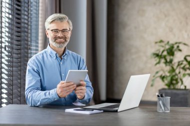 Modern bir ofis ortamında çalışırken gri saçlı ve gözlüklü gülümseyen kıdemli işadamı tablet kullanıyor. Bilgisayarı, defteri ve arka planda bitkisi olan bir masada oturuyor..