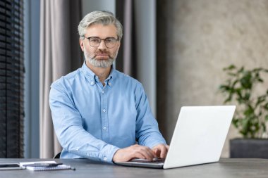 Modern ofis ortamında dizüstü bilgisayarda yazan gözlüklü, kendinden emin bir iş adamı. Görüntü profesyonelliği, odaklanmayı ve çağdaş bir çalışma ortamını betimliyor..