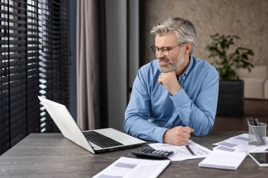 Modern ofis ortamındaki kağıtlarla dolu bir masada dizüstü bilgisayarda çalışan gözlüklü yaşlı bir adam. Gülümsüyor ve odaklanmış görünüyor. Bu da verimli ve olumlu bir çalışma ortamı olduğunu gösteriyor..