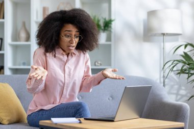 Kafası karışık bir kadın koltukta oturmuş, hayal kırıklığına uğramış bir ifadeyle evde laptopuyla çalışıyor. Görüntü, uzak çalışma zorlukları, duygusal stres ve sorun çözme zorluklarını betimliyor.
