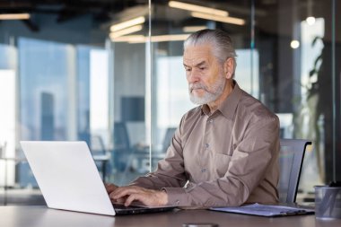 Modern bir ofiste dizüstü bilgisayar üzerinde çalışan kıdemli bir adam. Resim, profesyonel bir ortamda odak ve konsantrasyon, deneyim ve üretkenliği vurguluyor