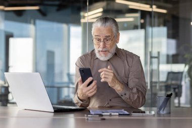 Üst düzey işadamı modern ofis ortamında akıllı telefon ve dizüstü bilgisayar kullanıyor. Üretkenliği ve etkili iletişimi gösteren profesyonel bir göreve odaklanmış görünüyor..