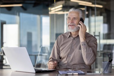 Üst düzey işadamı akıllı telefonla konuşurken gülümsüyor ve modern bir ofiste dizüstü bilgisayarla çalışıyor. İmaj, bir iş ortamında profesyonellik, teknoloji ve verimliliği resmediyor..