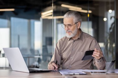 Üst düzey işadamı bir dizüstü bilgisayarda çalışıyor ve modern ofis ortamında tablet tutuyor. Profesyonel atmosfer üretkenlik, deneyim ve teknoloji işletmelerinin kullanımını sergiliyor.