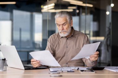 Çağdaş bir ofis ortamında dizüstü bilgisayar üzerinde çalışırken yaşlı bir erkek evrak işleriyle uğraşıyor. Adam belgelerin detaylarına odaklanıyor. Konsantrasyon ve çalışkanlığı yansıtıyor..