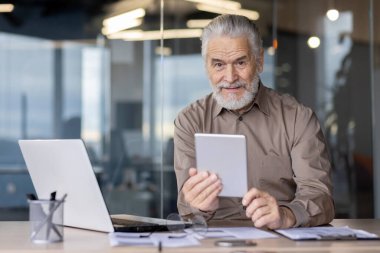 Yaşlı işadamı ofis masasında bir tablet kullanıyor, etrafı dizüstü bilgisayar ve belgelerle çevrili. Kıdemli adam kendinden emin bir şekilde gülümsüyor, deneyimi ve modern teknolojiyi profesyonel bir ortamda somutlaştırıyor..