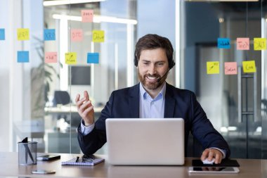 Modern ofis ortamında bir iş adamı, sanal bir toplantıda. Dizüstü bilgisayar kullanıyor ve kulaklık takıyor. Etrafı renkli notlarla çevrili. Fikirler ve planlarla dolu..
