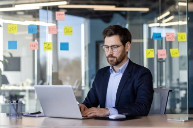 Takım elbiseli odaklanmış bir işadamı modern bir ofiste dizüstü bilgisayarıyla çalışıyor. Renkli yapışkan notları olan cam duvarlar çalışma alanına yaratıcı bir dokunuş katıyor. Konsept: İş, iş, meslek.