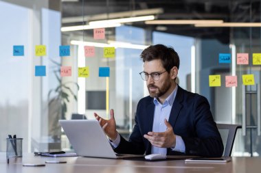 Modern bir ofiste profesyonel bir işadamı bir projeyi tartışırken video görüşmesi yapıyor. Etrafı cam duvardaki renkli yapışkan notlarla çevrili. İşbirlikçi ve organize olduğunu gösteriyor.