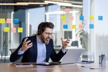 Kulaklık takan kızgın bir patron, modern bir ofisteki iş görüşmesi sırasında bir video görüşmesinde bağırıyor. Arkaplanda cam duvarlar ve ofis ekipmanları üzerine yapışkan notlar yer alıyor.