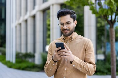 Ofis binasının dışında akıllı telefon kullanan genç işadamı, cihaza odaklanmış. Günlük kıyafetler, modern şirket ortamı, profesyonel yaşam tarzı. Şehir dışı, bağlı.