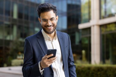 Modern ofis binasının dışında akıllı telefona bakarken resmi takım elbiseli bir işadamı gülümsüyor. İş, meslek, iletişim, teknoloji ve kurumsal yaşam tarzı kavramı
