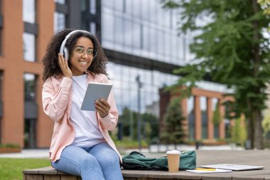 Üniversite kampüsünde dışarıda otururken dijital tablet ve kulaklık kullanan gülümseyen genç bir kadın. Müzikten ve teknolojiden zevk alıyor, günlük giysiler giyiyor. Güneşli bir günde rahat ve mutlu titreşimler..