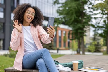 Şehir parkındaki video görüşmesi sırasında bankta oturmuş akıllı telefona el sallayan gülümseyen genç bir kadın. Güneşli bir günde kahve, kulaklık ve tabletle keyifli sohbetler..