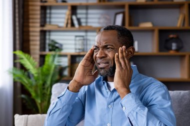 Modern oturma odasındaki koltukta oturan, başı ağrıyan stresli adam. Kafasını rahatsızlık içinde tutuyor, acı ve endişeye işaret ediyor. Kitap rafı ve bitkilerle dolu parlak, iyi organize edilmiş bir arka plan..