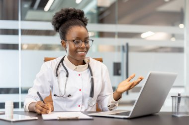 Beyaz önlük giyen gülümseyen doktor ve steteskop modern ofiste video aracılığıyla sanal konsültasyon yapıyor. Dijital platform vasıtasıyla hastaya profesyonel tıbbi danışma.