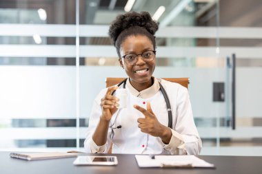 Online konsültasyon sırasında elinde reçete şişesiyle gülümseyen kadın doktor. Hastayla görüntülü görüşme yaparak ilgilenmek, ilaçları işaret etmek. Modern ofis ayarları, masadaki tablet belgeleri.