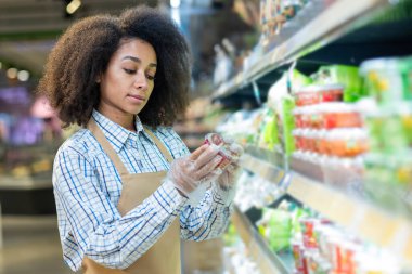 Süpermarket çalışanı marketteki raflardaki ürün etiketlerini kontrol ediyor. Önlük takan ve yiyecek paketlerini inceleyen eldiven takan kadın perakende işçisi. Perakende satış, market kalite kontrolü kavramı