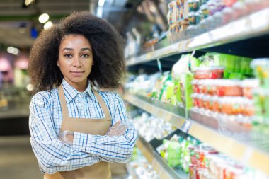 Önlüklü, mağaza koridorunda kolları çapraz duran kendinden emin bir market çalışanı. Raflar arka planda çeşitli ürünlerle dolu. Perakende işi kavramı, müşteri hizmetleri