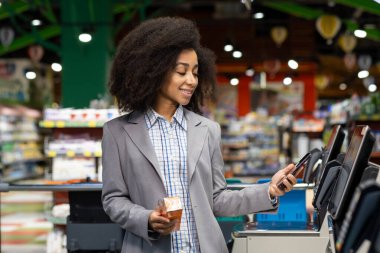 Marketteki kasiyerde akıllı telefon kullanan gülümseyen bir kadın. Profesyonel kıyafet, elinde içecek bardağı var. Modern alışveriş deneyimi, kolaylık, teknoloji
