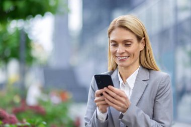 Açık havada akıllı telefon kullanırken gülümseyen gri takım elbiseli kendine güvenen bir iş kadını. Profesyonel kadın şehir ortamında mobil cihazlar aracılığıyla iletişim ve ağ kuruyor..