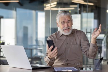 Yaşlı işadamı akıllı telefon tutarken ve dizüstü bilgisayara bakarken kafa karışıklığı yaşıyor. Modern ofiste cam duvarlı bir sahne. Teknoloji sorunları kavramı, kıdemli işçi adaptasyonu