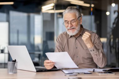 Üst düzey işadamı, modern ofiste dizüstü bilgisayardaki belgeleri incelerken başarıyı heyecanla kutluyor. Profesyonel zevkle yumruğunu sıkıp başarıyı kutluyor..