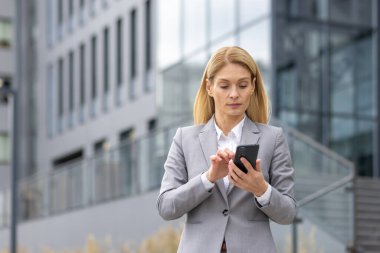 Modern ofis binasının dışında akıllı telefon kullanan gri takım elbiseli bir iş kadını. Profesyonel ve odaklanmış kadın mesajları kontrol ediyor veya dijital aygıt üzerinde çalışıyor.