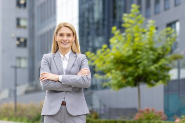Gri takım elbiseli kendine güvenen iş kadını şehir ortamında kollarını kavuşturup gülümsüyor. Arka planda modern binalar ve yeşillik var. Başarı, güven, liderlik ve iş kavramı.