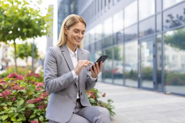 Gri takım elbiseli iş kadını ofis binasında akıllı telefon kullanıyor. Profesyonel bir kadın gülümsüyor ve teknolojiyle etkileşimde bulunuyor. Cam bina ve yeşillik ile modern kentsel ortam.