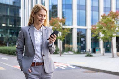 Gri takım elbiseli iş kadını modern ofis binasının dışında akıllı telefon kullanıyor. Cep telefonu tutarken gülümseyen profesyonel bir kadın, başarılı iş hayatı tarzını canlandırıyor..