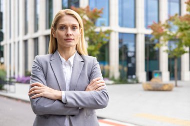Modern ofis binasının dışında kolları bağlı duran gri takım elbiseli kendine güvenen bir iş kadını. Profesyonel ve kararlı bir ifade. Açık şehir ortamında başarılı bir şirket kadını..
