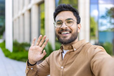 Gözlüklü ve sıradan giyinmiş mutlu genç bir adam açık hava video görüşmesi sırasında el sallıyor ve gülümsüyor. Yeşillik içinde modern binanın önünde dikilen adam neşe, dostluk ve bağlılık ifade ediyor..