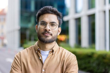 Gözlüklü ve sakallı genç adam şehir ortamında güvenle ayakta duruyor. Gündelik kıyafetler ve modern arka plan, çağdaş şehir yaşamını ve mesleki tutumunu vurguluyor.