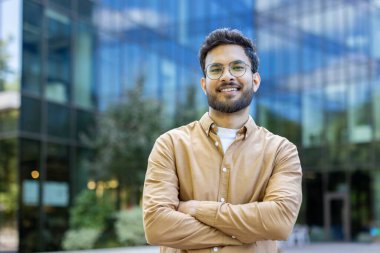 Gözlüklü genç profesyonel adam modern cam ofis binasının önünde güvenle duruyor, gülümsüyor ve kollarını kavuşturuyor. Kentsel çevrede iş ve başarı kavramı.