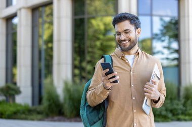 Modern binanın önünde duran üniversite öğrencisi telefona bakarken gülümsüyor. Kampüste eğitim, teknoloji ve gençlik yaşam tarzını temsil eden sırt çantası ve klasör taşıyan öğrenci.